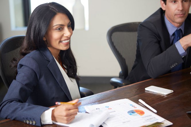 A smiling woman in a suit, sitting as a table in a conference room.