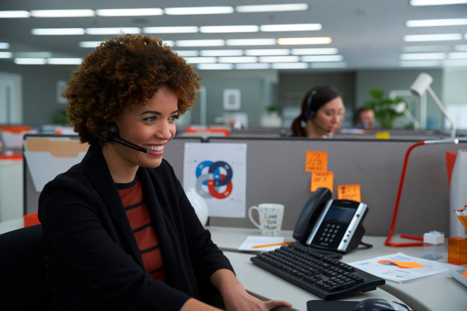 A smiling woman in an office, using a headset to speak on the telephone.