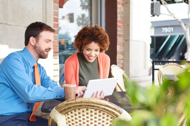 A man and a woman sitting outside at a coffee shop, using a tablet.