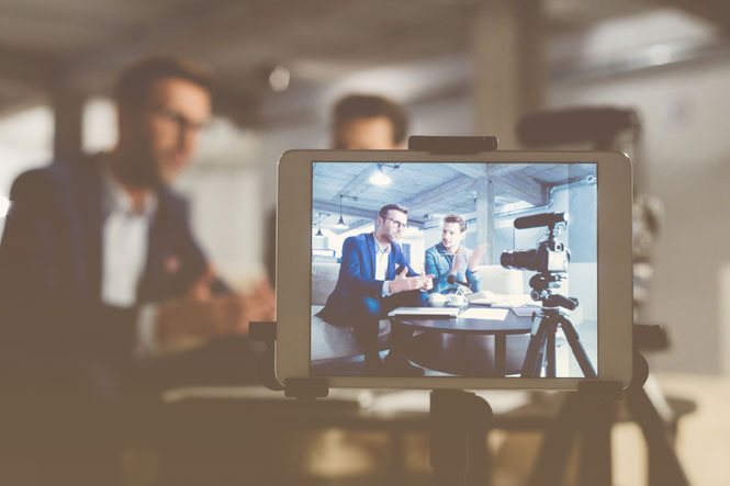 Two professionals talk to colleagues via a video conference app while a camera records them