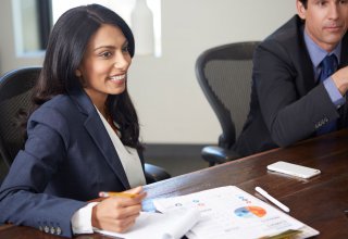 A smiling woman in a suit, sitting as a table in a conference room.