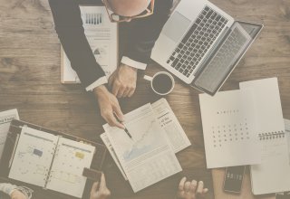 Person reviewing budget planning with two others, using a laptop and charts on paper