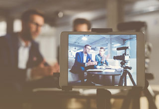 Two professionals talk to colleagues via a video conference app while a camera records them